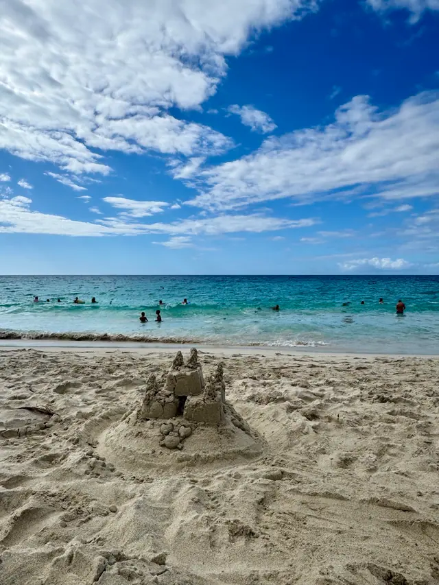 A sandcastle on a white-sand beach with swimmers in the turquoise sea beyond