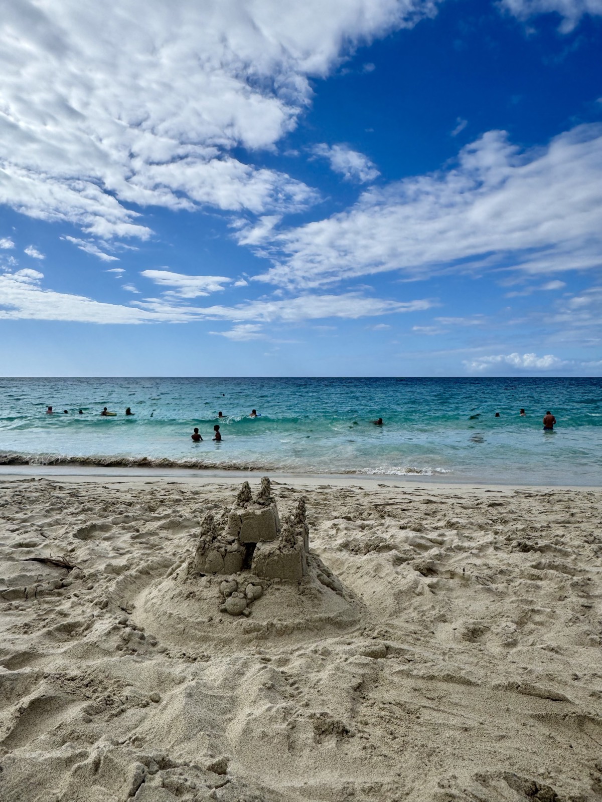 A sandcastle on a white-sand beach with swimmers in the turquoise sea beyond