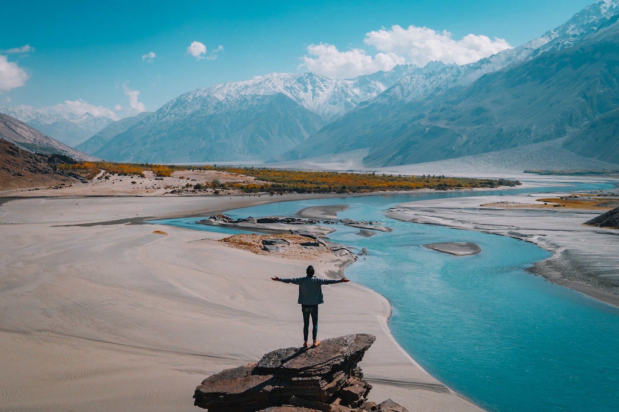 A traveler with arms outstretched above a turquoise river valley beneath snow-capped mountains